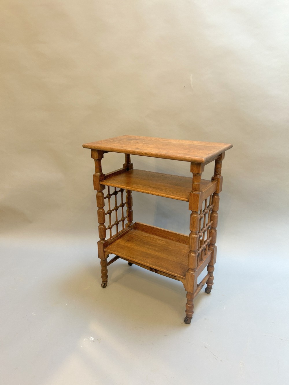 An Arts and Crafts oak side table featuring three tiers, turned spindle supports, open lattice side panels, and brass castors.