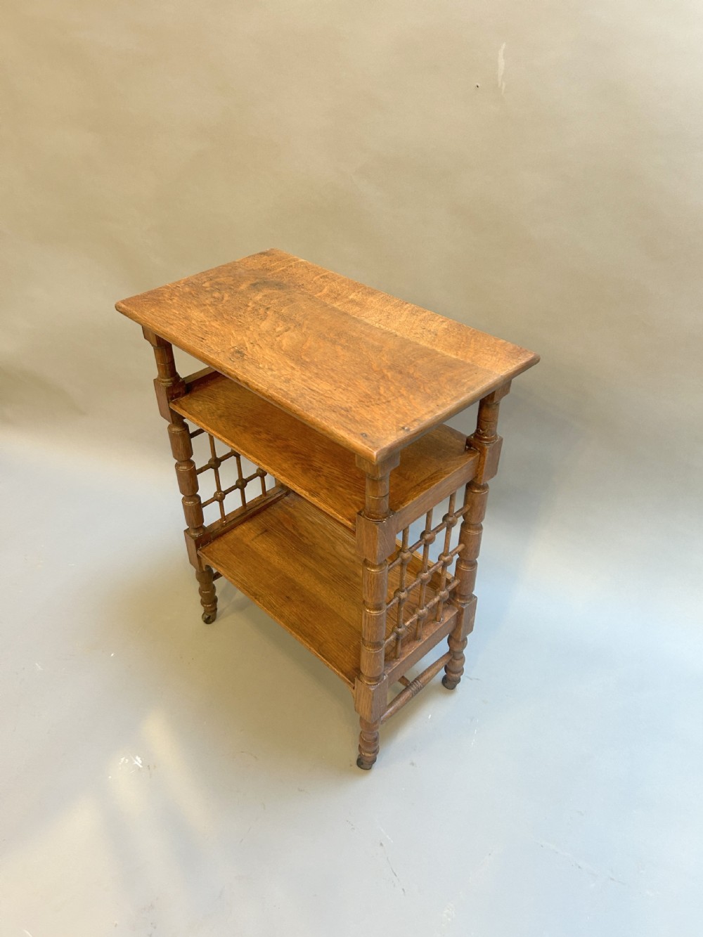 An Arts and Crafts oak side table featuring three tiers, turned spindle supports, open lattice side panels, and brass castors. View II