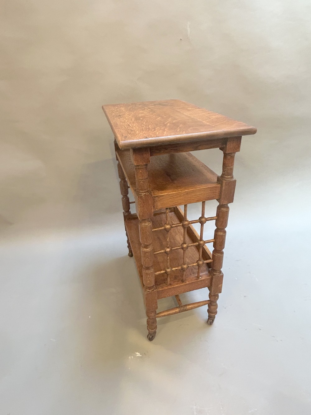 An Arts and Crafts oak side table featuring three tiers, turned spindle supports, open lattice side panels, and brass castors. View III