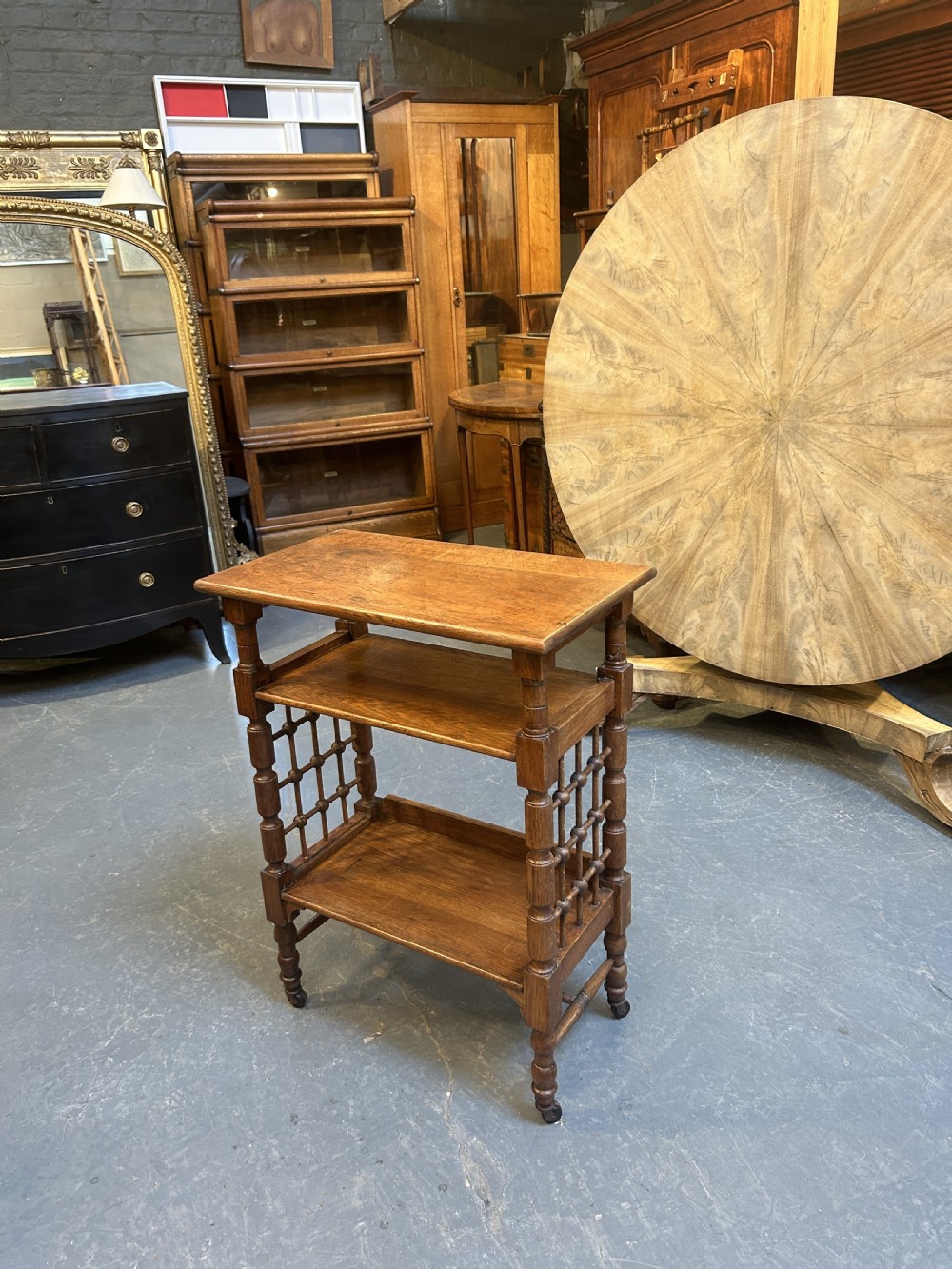 An Arts and Crafts oak side table featuring three tiers, turned spindle supports, open lattice side panels, and brass castors. View IV