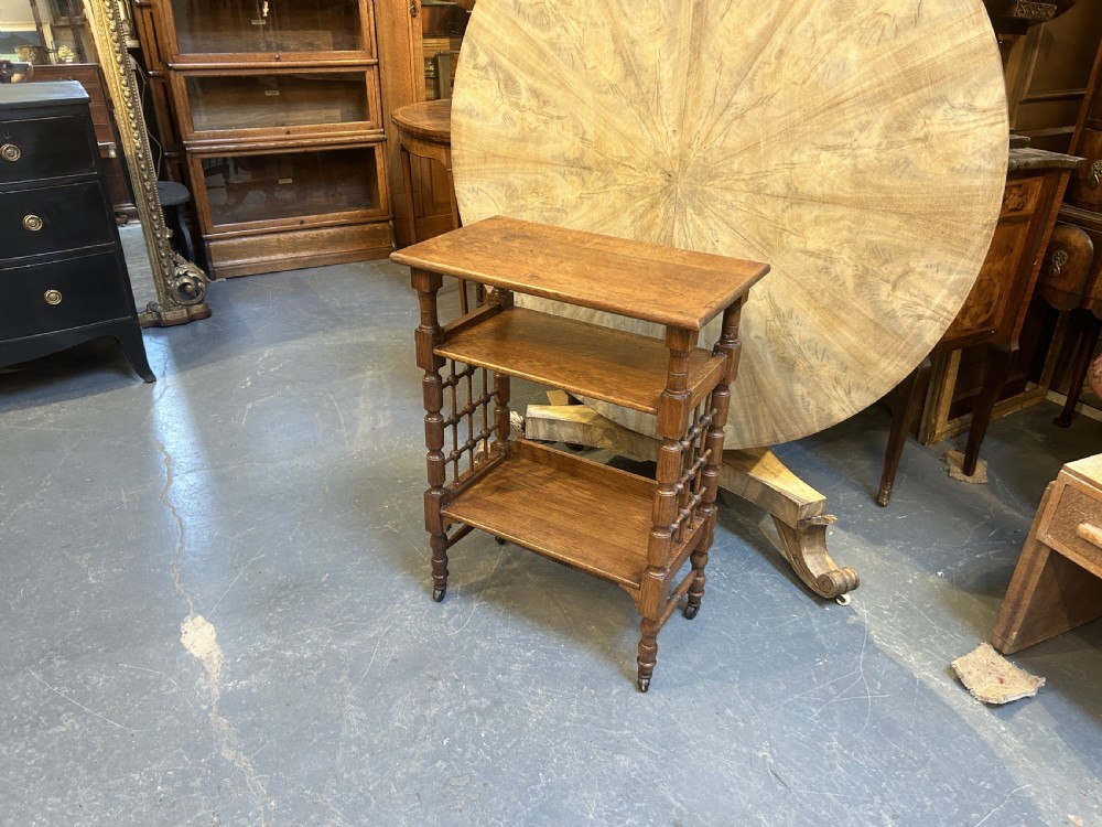 An Arts and Crafts oak side table featuring three tiers, turned spindle supports, open lattice side panels, and brass castors. View V