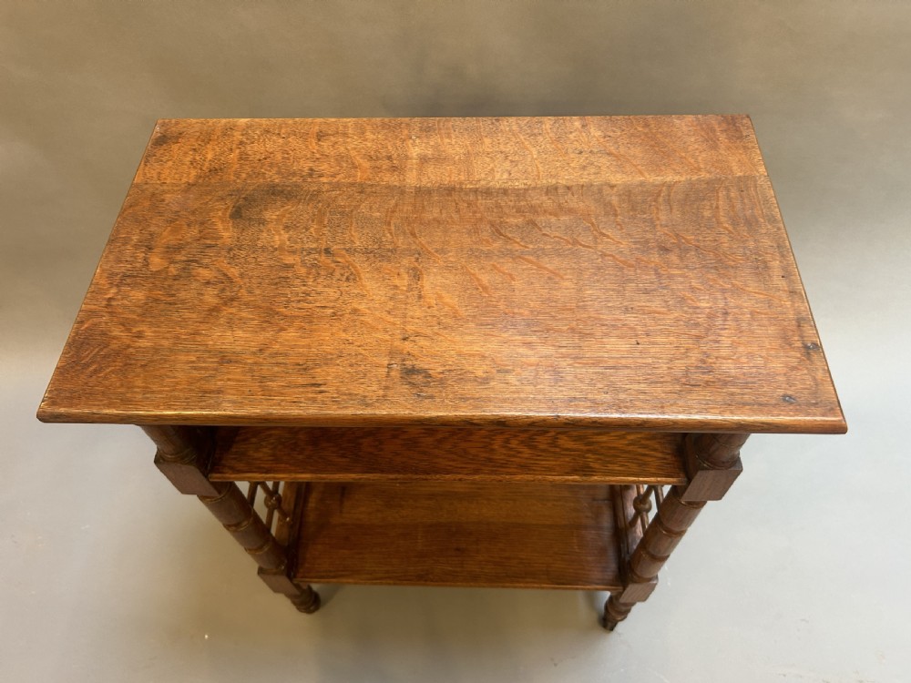An Arts and Crafts oak side table featuring three tiers, turned spindle supports, open lattice side panels, and brass castors. View VI