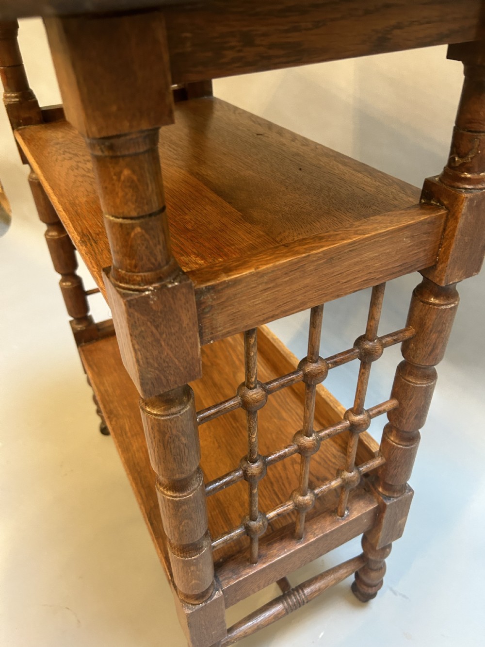An Arts and Crafts oak side table featuring three tiers, turned spindle supports, open lattice side panels, and brass castors. View VII