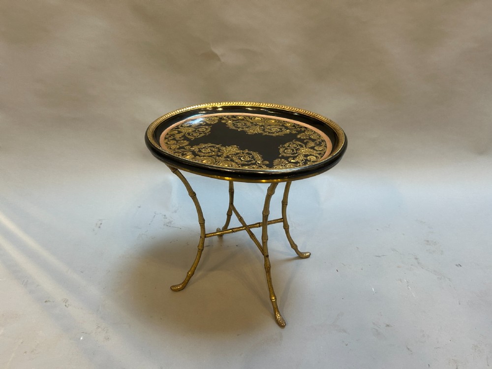 An oval black lacquered papier-mache tray table with detailed gilt scrollwork, supported on a brass faux-bamboo base with an X-stretcher.