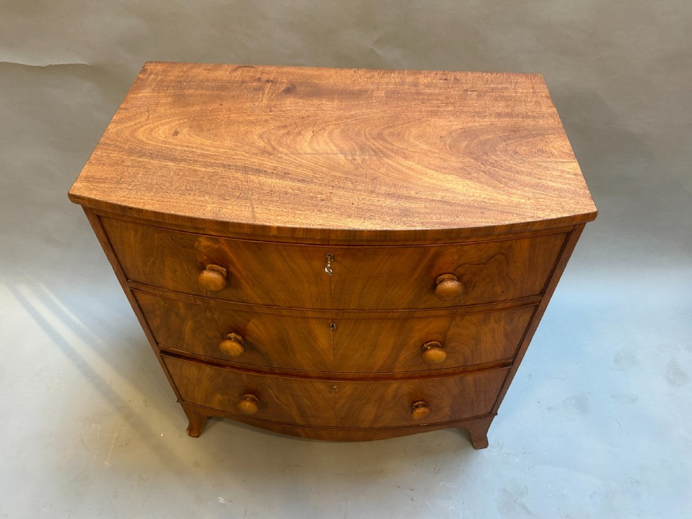 A George III bow front chest of drawers in warm brown timber, featuring three graduated drawers with turned wooden handles and splayed feet. View V