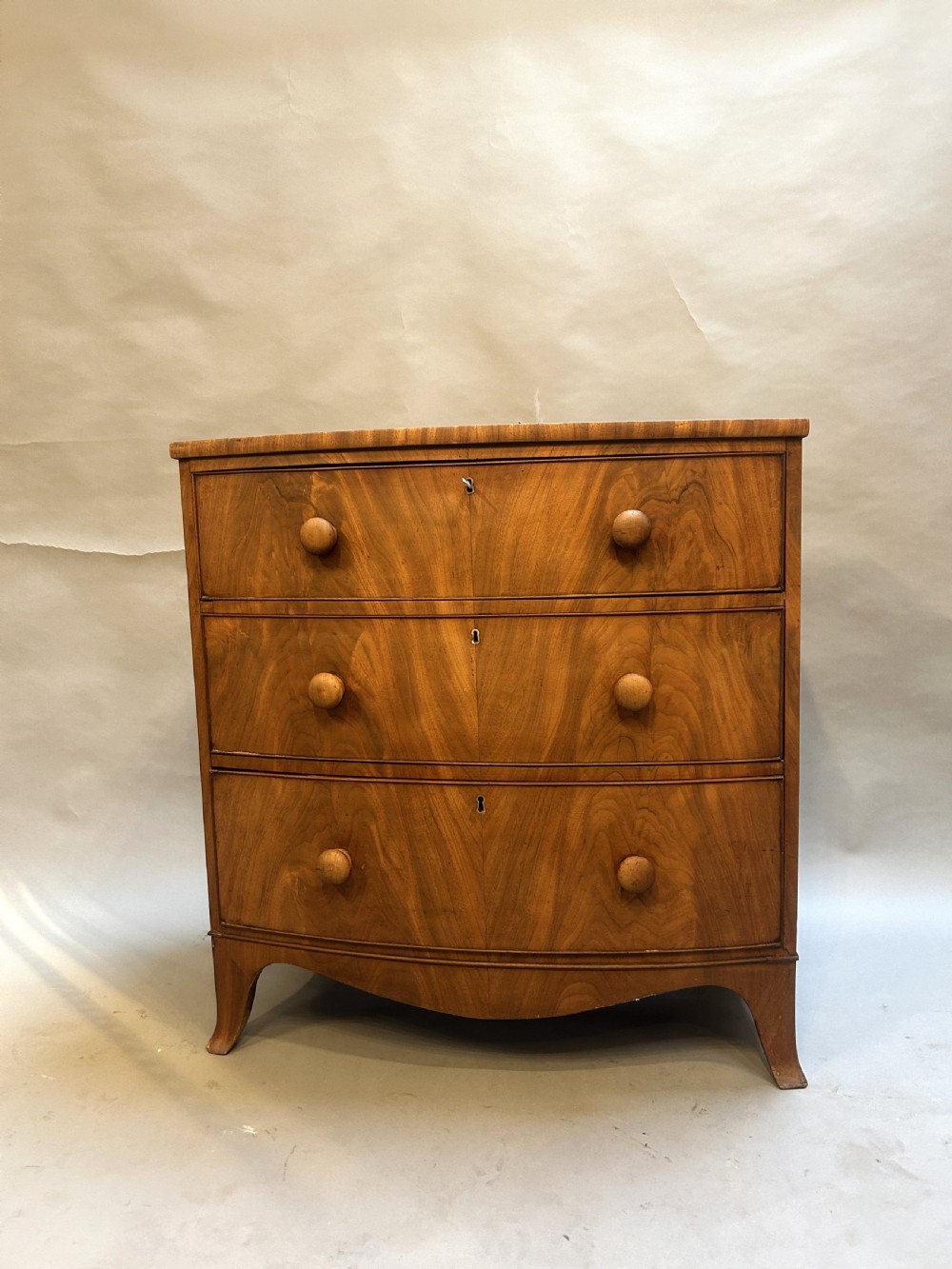 A George III bow front chest of drawers in warm brown timber, featuring three graduated drawers with turned wooden handles and splayed feet. View VI