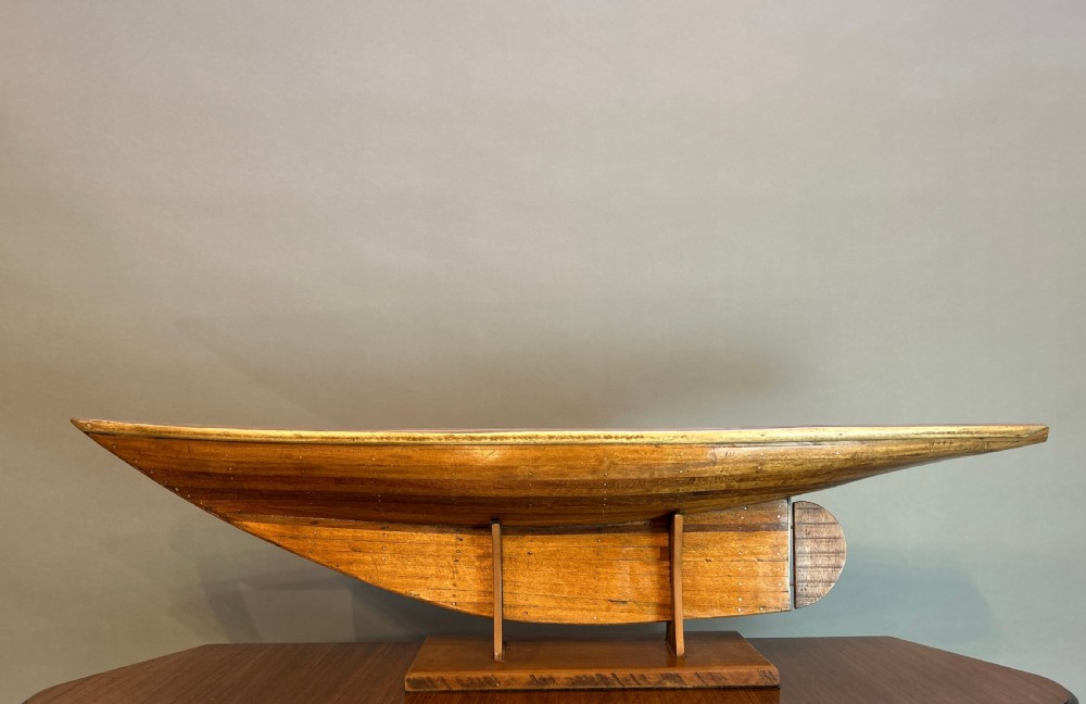 A varnished wooden pond yacht hull supported upright on a custom wooden display stand.