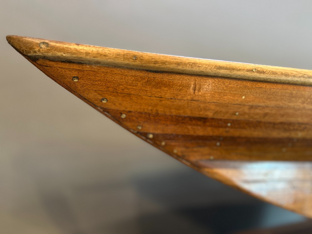 A varnished wooden pond yacht hull supported upright on a custom wooden display stand. View IV