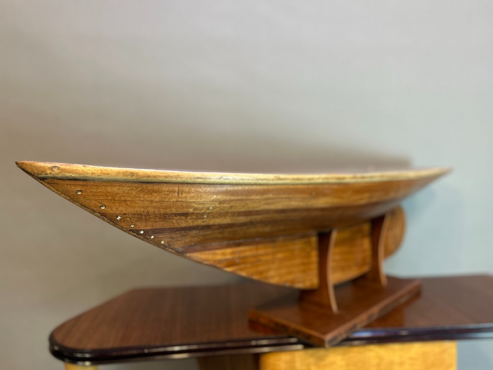 A varnished wooden pond yacht hull supported upright on a custom wooden display stand. View V