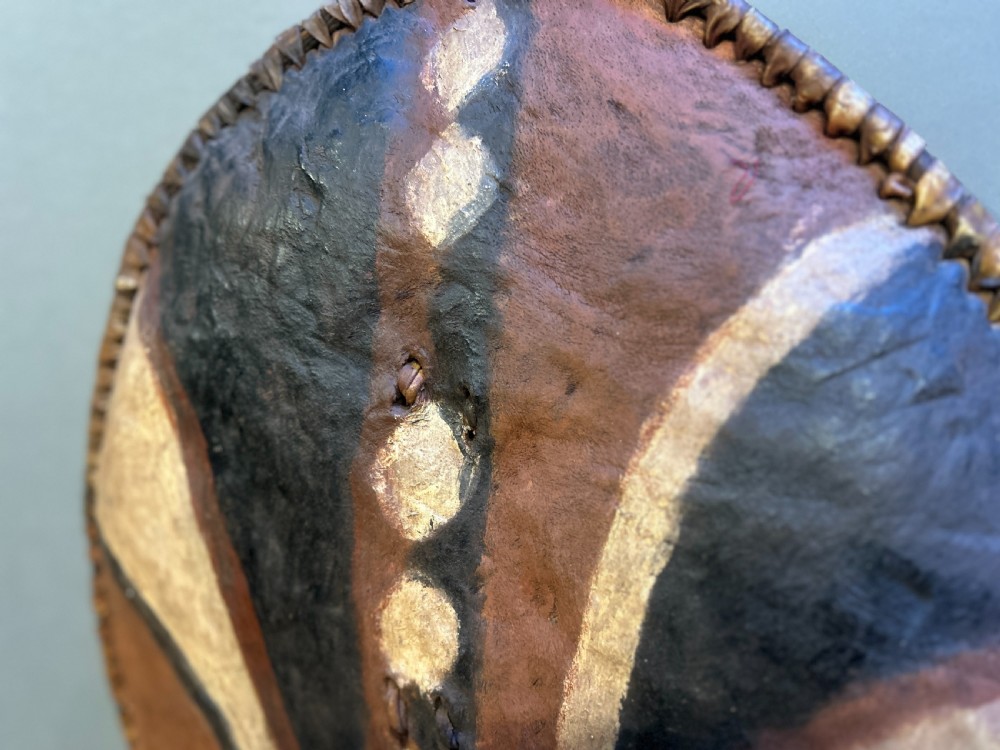 An oval African hide shield featuring hand-painted black, brown, and ochre geometric patterns with stitched edge detailing. View II
