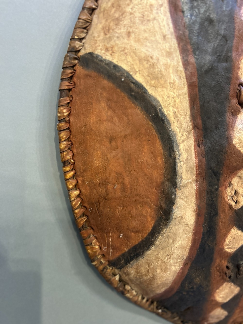 An oval African hide shield featuring hand-painted black, brown, and ochre geometric patterns with stitched edge detailing. View III