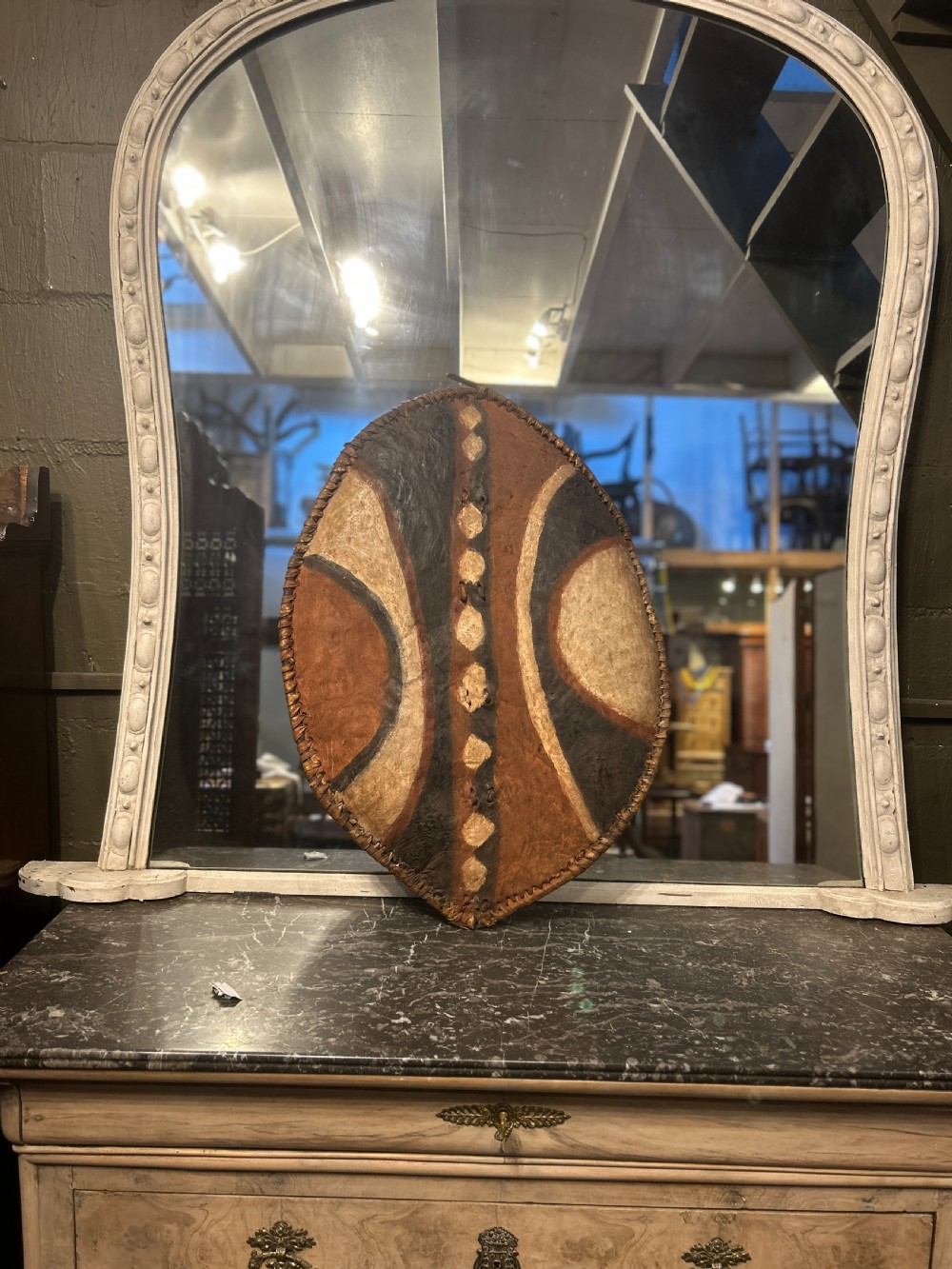 An oval African hide shield featuring hand-painted black, brown, and ochre geometric patterns with stitched edge detailing. View IV