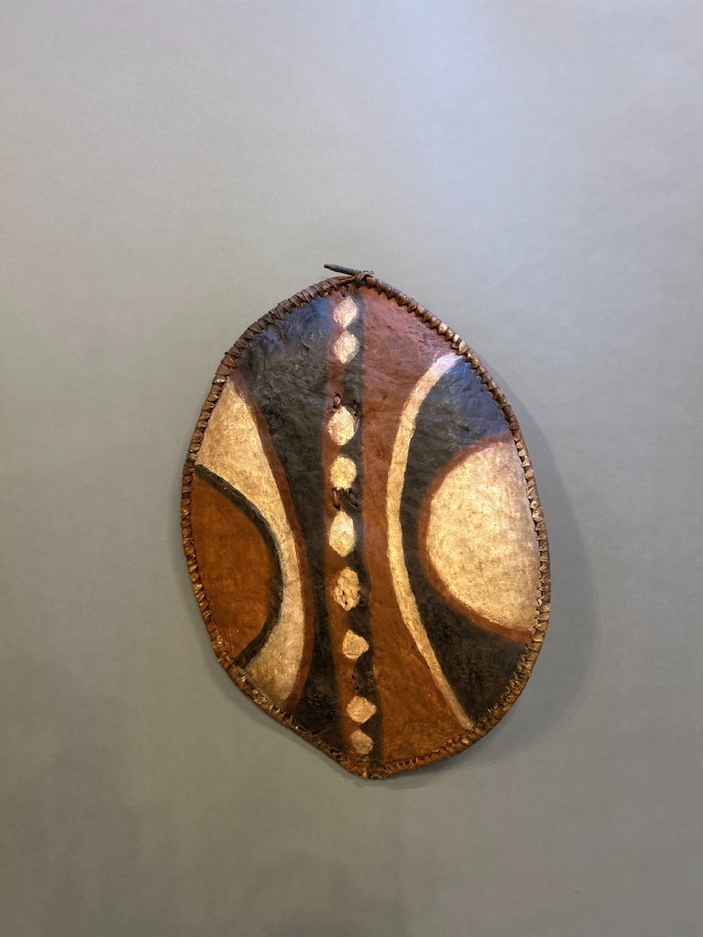 An oval African hide shield featuring hand-painted black, brown, and ochre geometric patterns with stitched edge detailing.