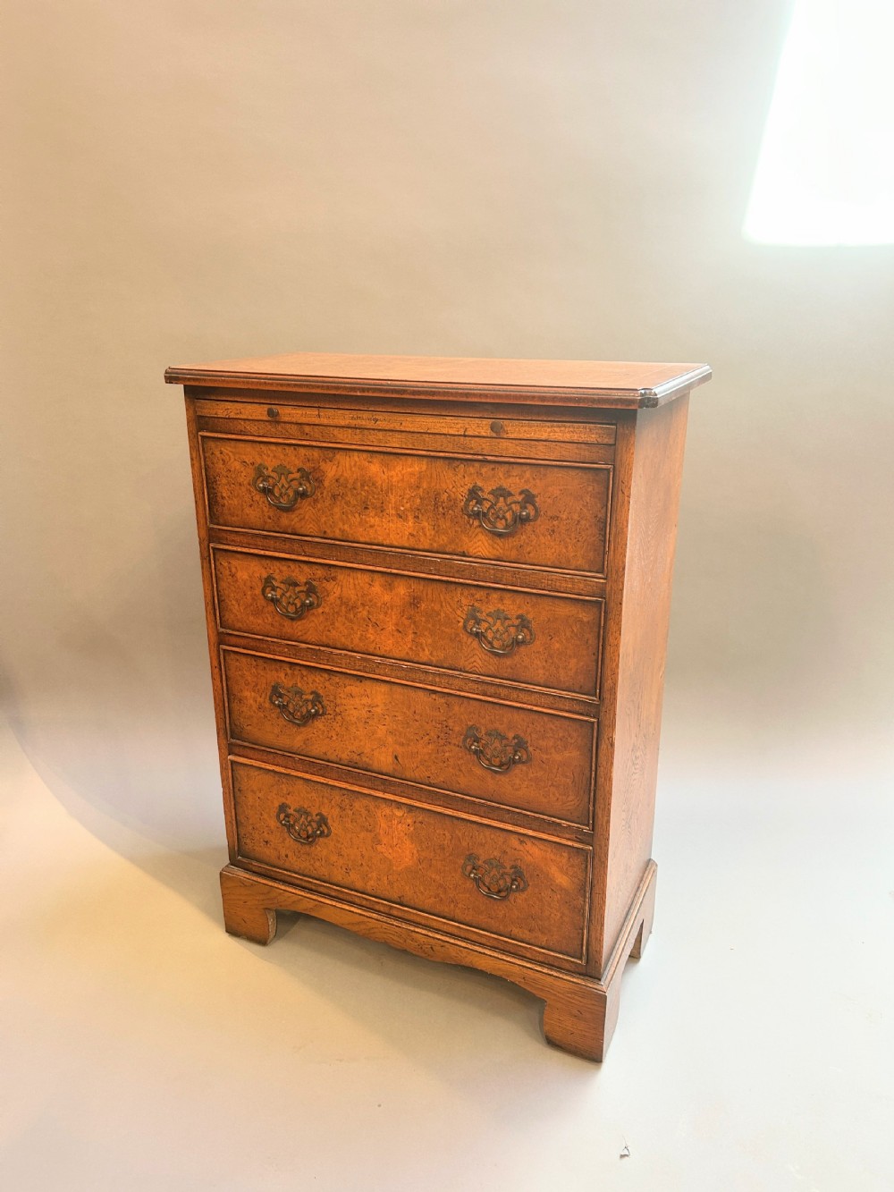 A small Georgian Revival burr walnut chest of drawers featuring four graduated drawers with brass drop handles and bracket feet.