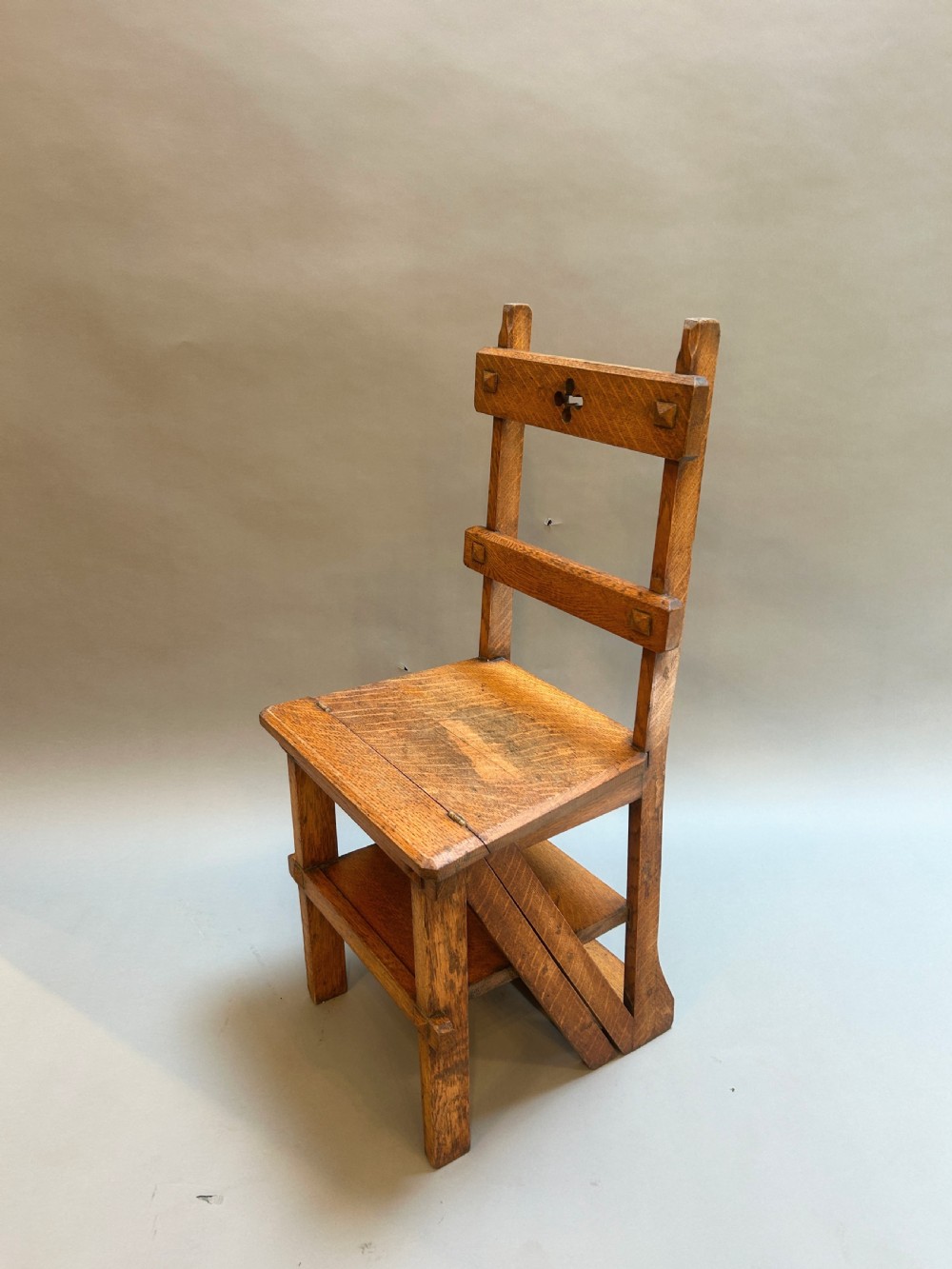 An antique solid oak metamorphic chair that folds into library steps, featuring a pierced trefoil motif backrest and square legs.