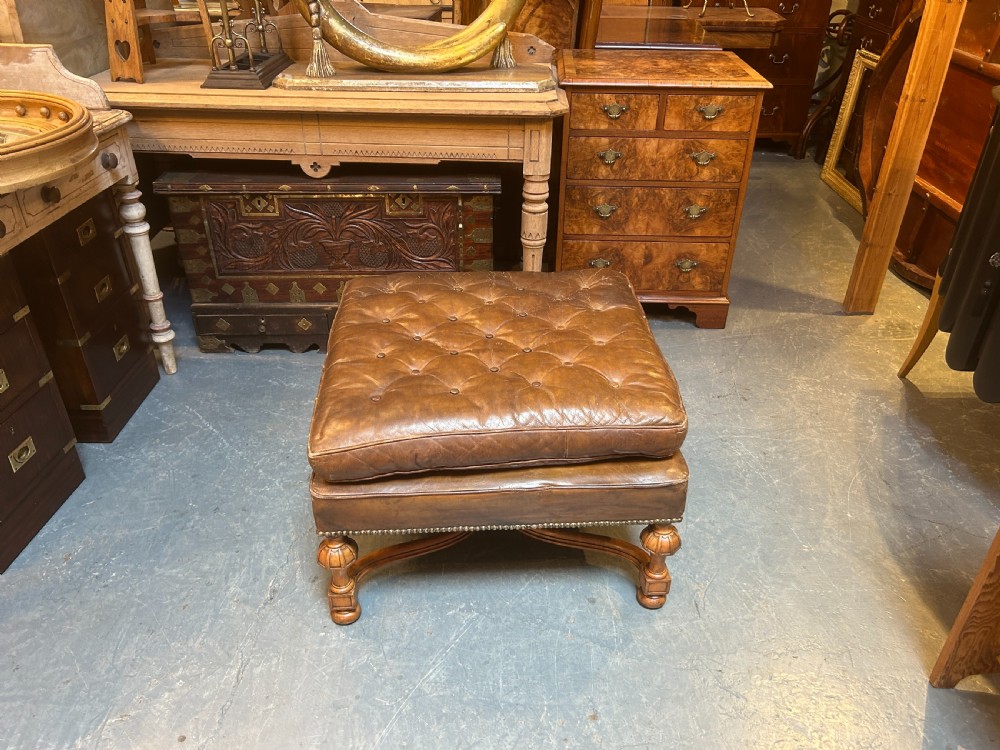 A square chestnut brown leather footstool featuring deep button tufting, brass stud trimming, and carved wooden legs with curved stretchers. View VI