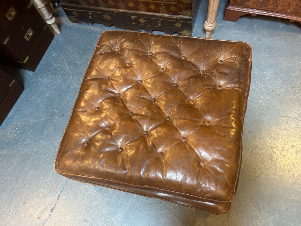 A square chestnut brown leather footstool featuring deep button tufting, brass stud trimming, and carved wooden legs with curved stretchers. View VII