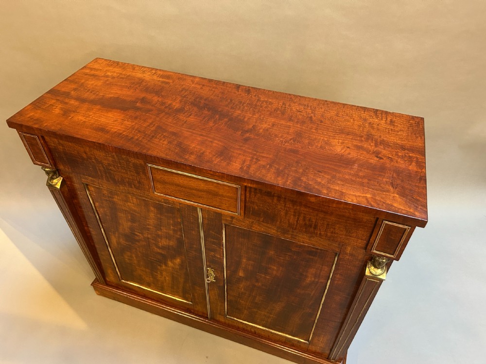 A Regency mahogany side cabinet featuring flame veneer, two panelled doors, brass stringing inlay, and gilt metal Egyptian bust mounts. View II