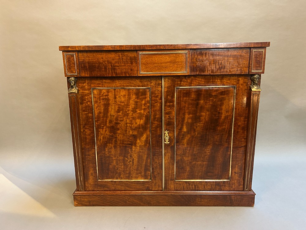 A Regency mahogany side cabinet featuring flame veneer, two panelled doors, brass stringing inlay, and gilt metal Egyptian bust mounts. View V