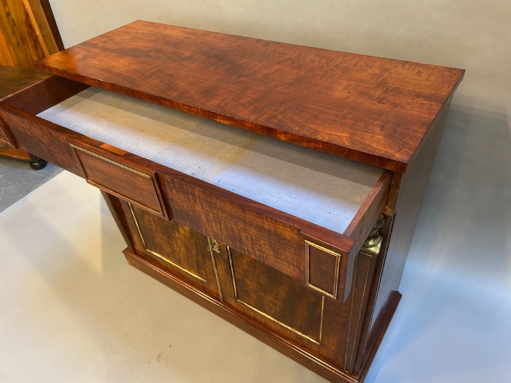 A Regency mahogany side cabinet featuring flame veneer, two panelled doors, brass stringing inlay, and gilt metal Egyptian bust mounts. View VII