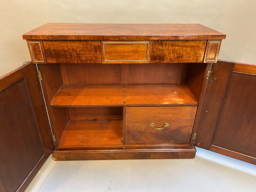 A Regency mahogany side cabinet featuring flame veneer, two panelled doors, brass stringing inlay, and gilt metal Egyptian bust mounts. View VIII