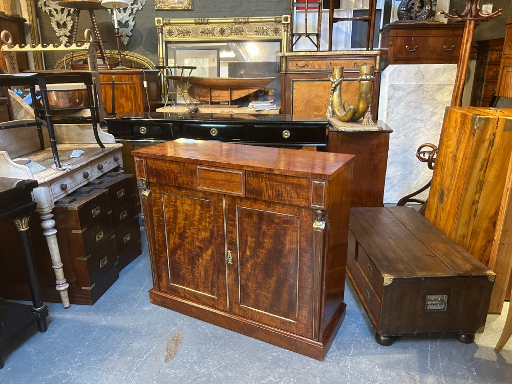 A Regency mahogany side cabinet featuring flame veneer, two panelled doors, brass stringing inlay, and gilt metal Egyptian bust mounts. View XIII