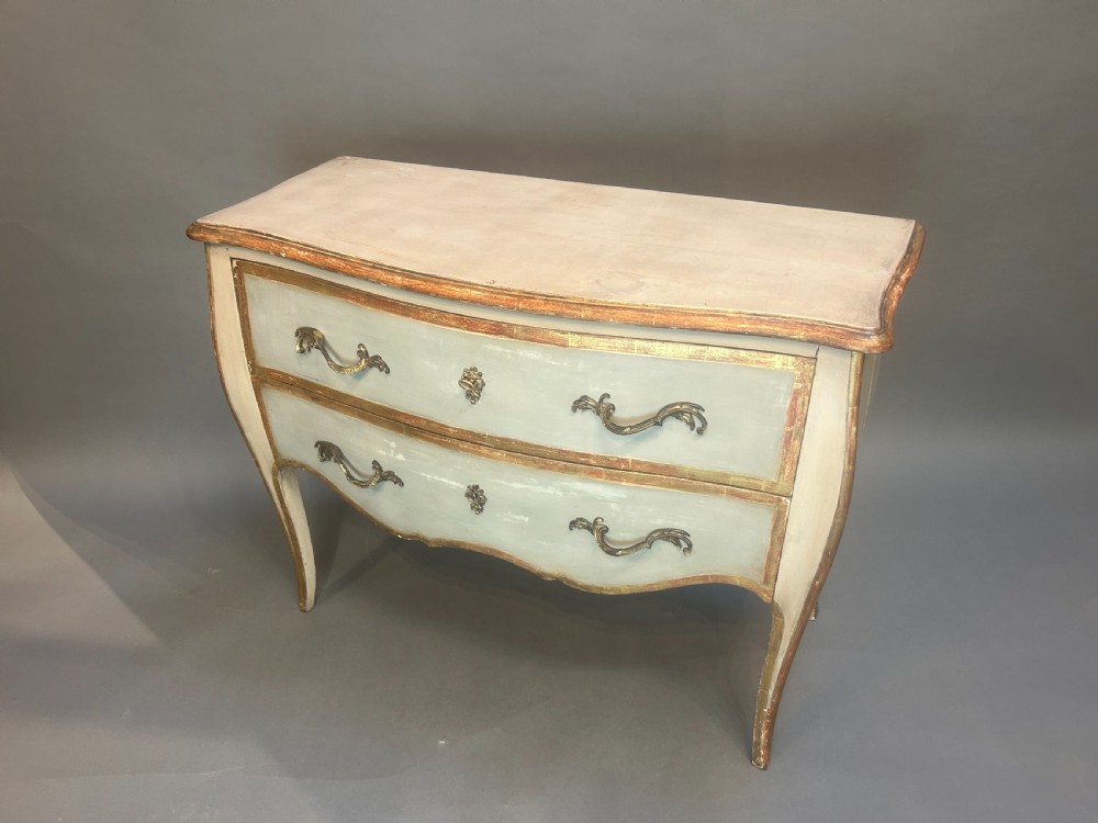 A French Louis XV style painted and gilt serpentine commode featuring two drawers, gold leaf borders, and curved cabriole legs.