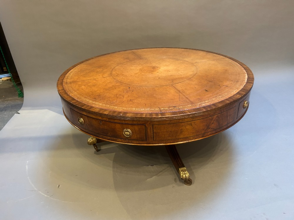A low circular mahogany drum table featuring a gold-tooled leather top, frieze drawers with brass pulls, and a pedestal base with brass lion paw castors.