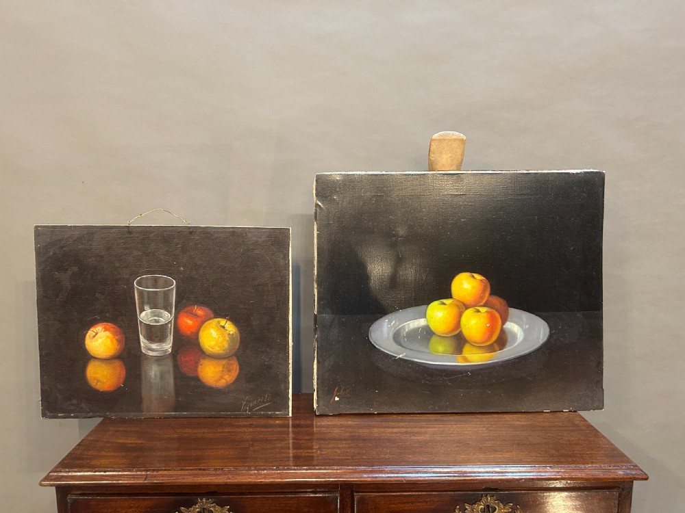 A pair of Spanish still-life oil paintings depicting apples, a pewter plate, and a glass of water against a dark background.