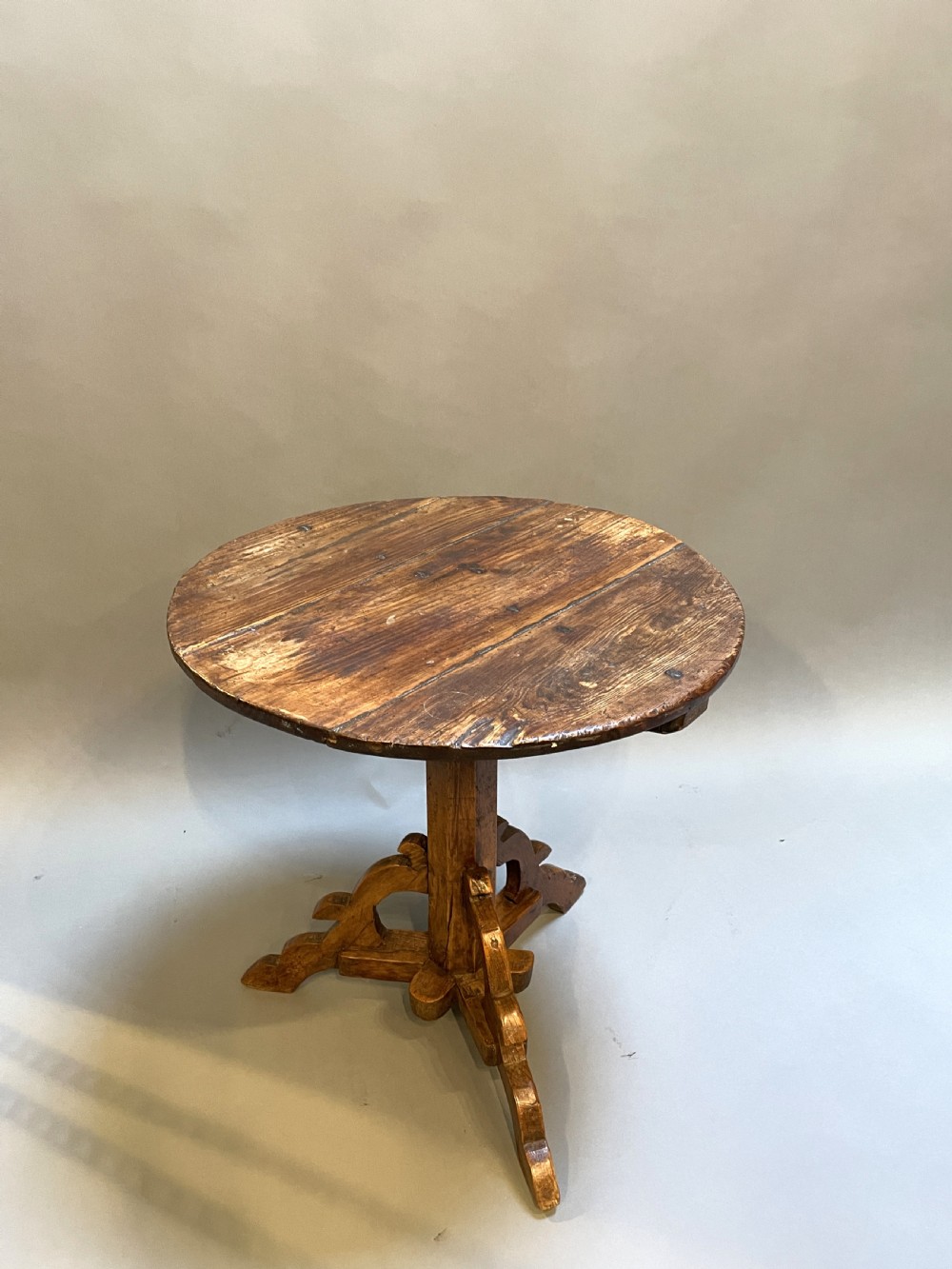 An 18th-century rustic pine pedestal table featuring a round plank top and carved tripod legs with a warm timber patina.