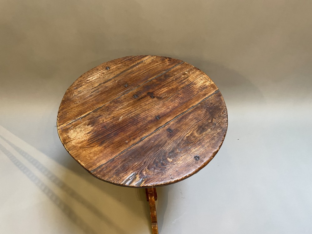 An 18th-century rustic pine pedestal table featuring a round plank top and carved tripod legs with a warm timber patina. View X