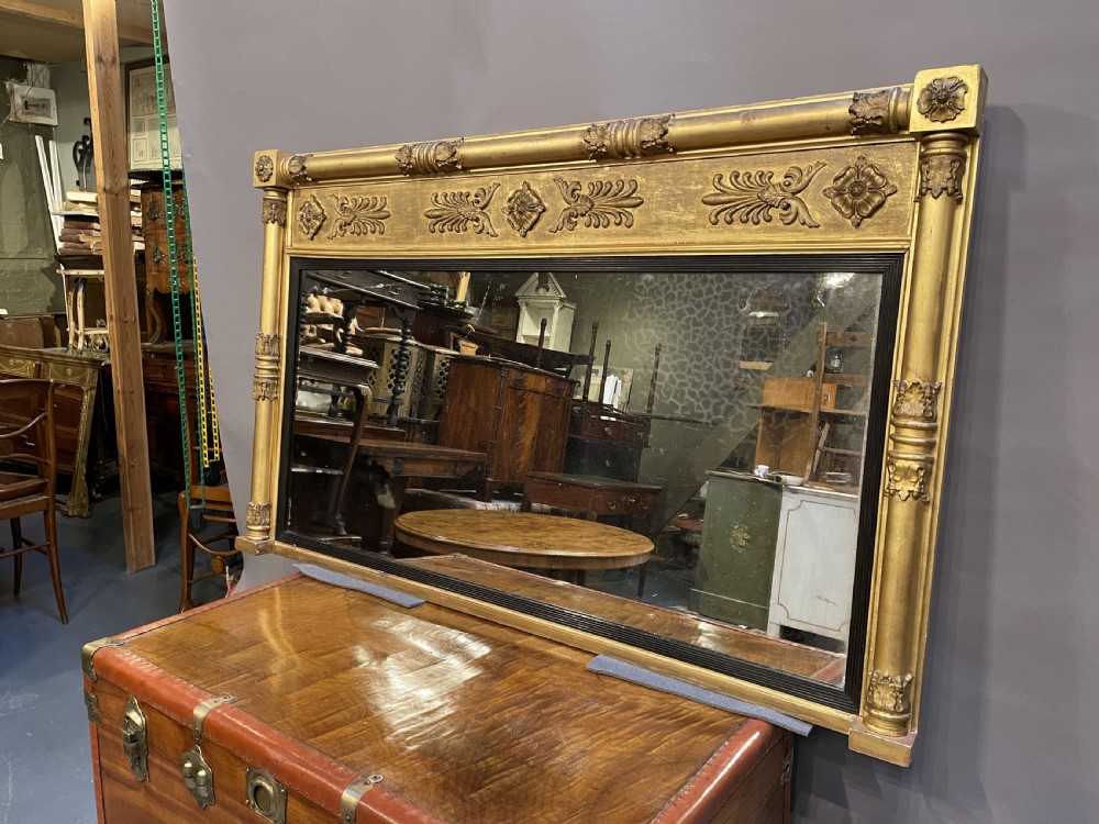 A large horizontal Regency gilt overmantel mirror featuring neoclassical side columns, a frieze with carved palmettes and rosettes, and an ebonised inner slip. View XIV