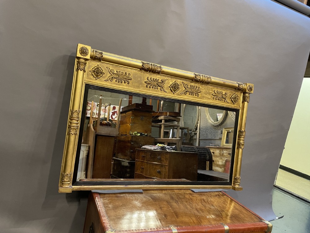 A large horizontal Regency gilt overmantel mirror featuring neoclassical side columns, a frieze with carved palmettes and rosettes, and an ebonised inner slip. View XV