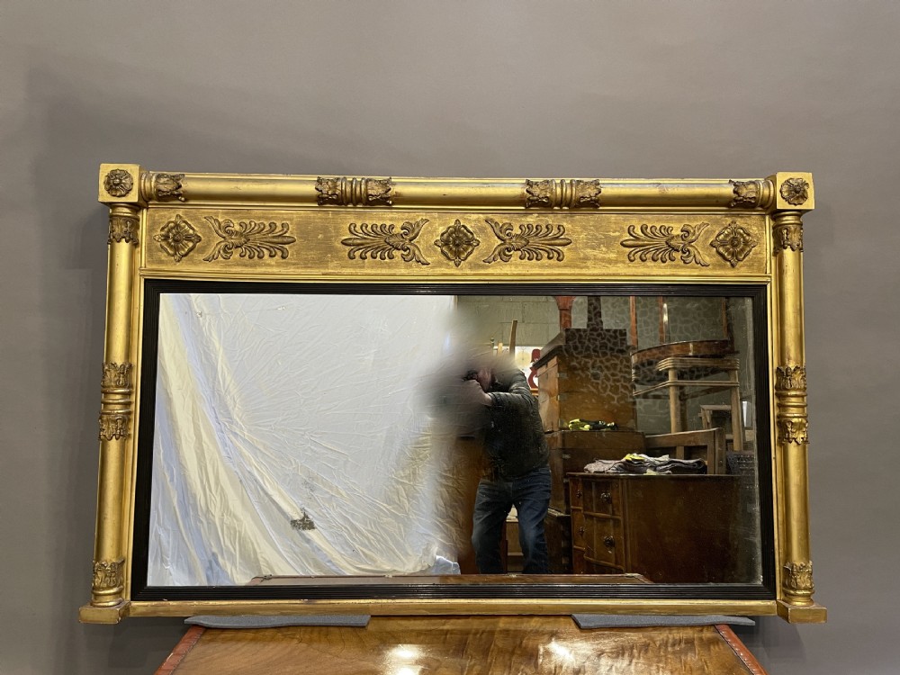 A large horizontal Regency gilt overmantel mirror featuring neoclassical side columns, a frieze with carved palmettes and rosettes, and an ebonised inner slip. View XVI