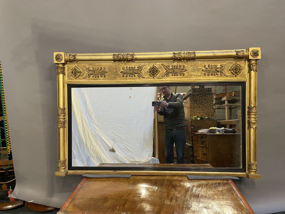 A large horizontal Regency gilt overmantel mirror featuring neoclassical side columns, a frieze with carved palmettes and rosettes, and an ebonised inner slip. View XVII