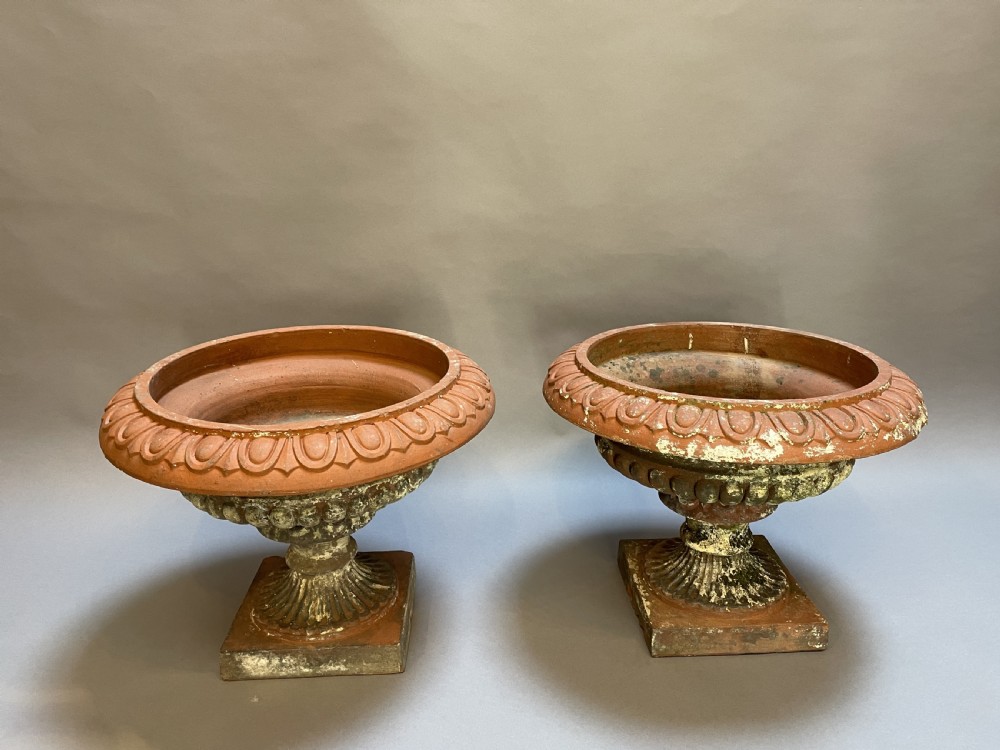 A pair of weathered terracotta Campana garden urns featuring wide egg-and-dart rims, gadrooned lower bowls, fluted stems, and square bases.