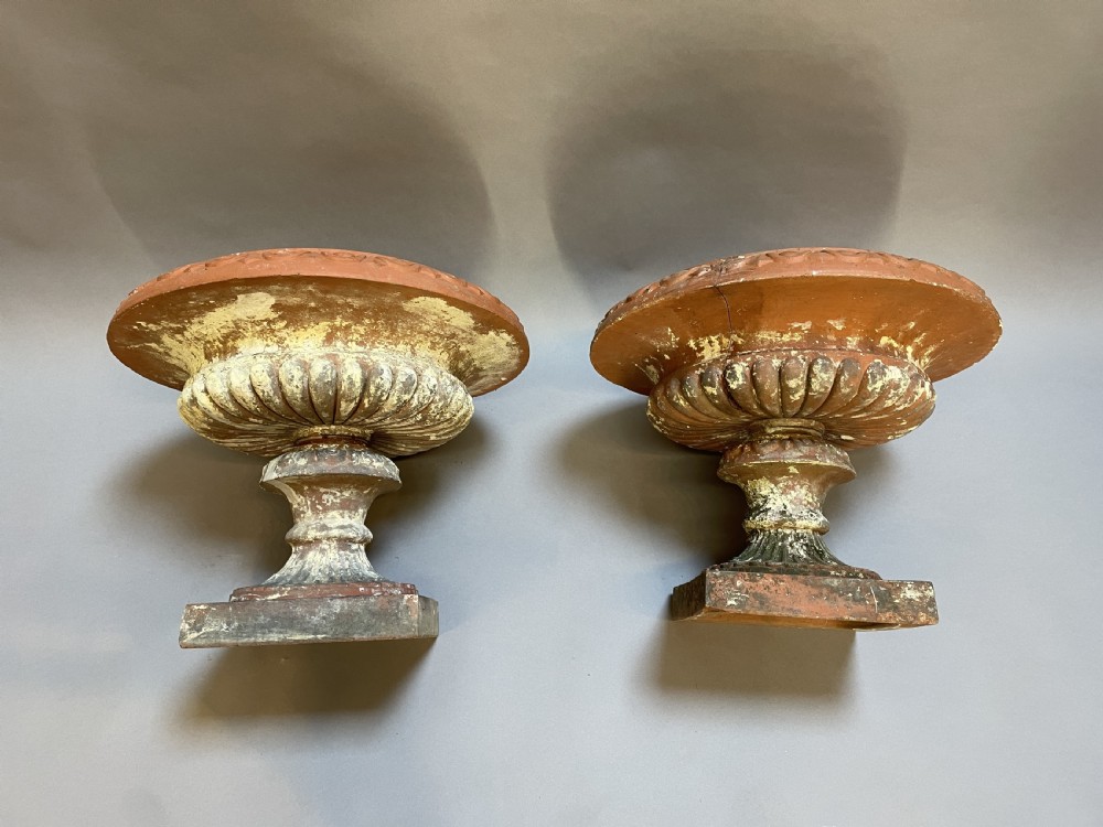 A pair of weathered terracotta Campana garden urns featuring wide egg-and-dart rims, gadrooned lower bowls, fluted stems, and square bases. View IX