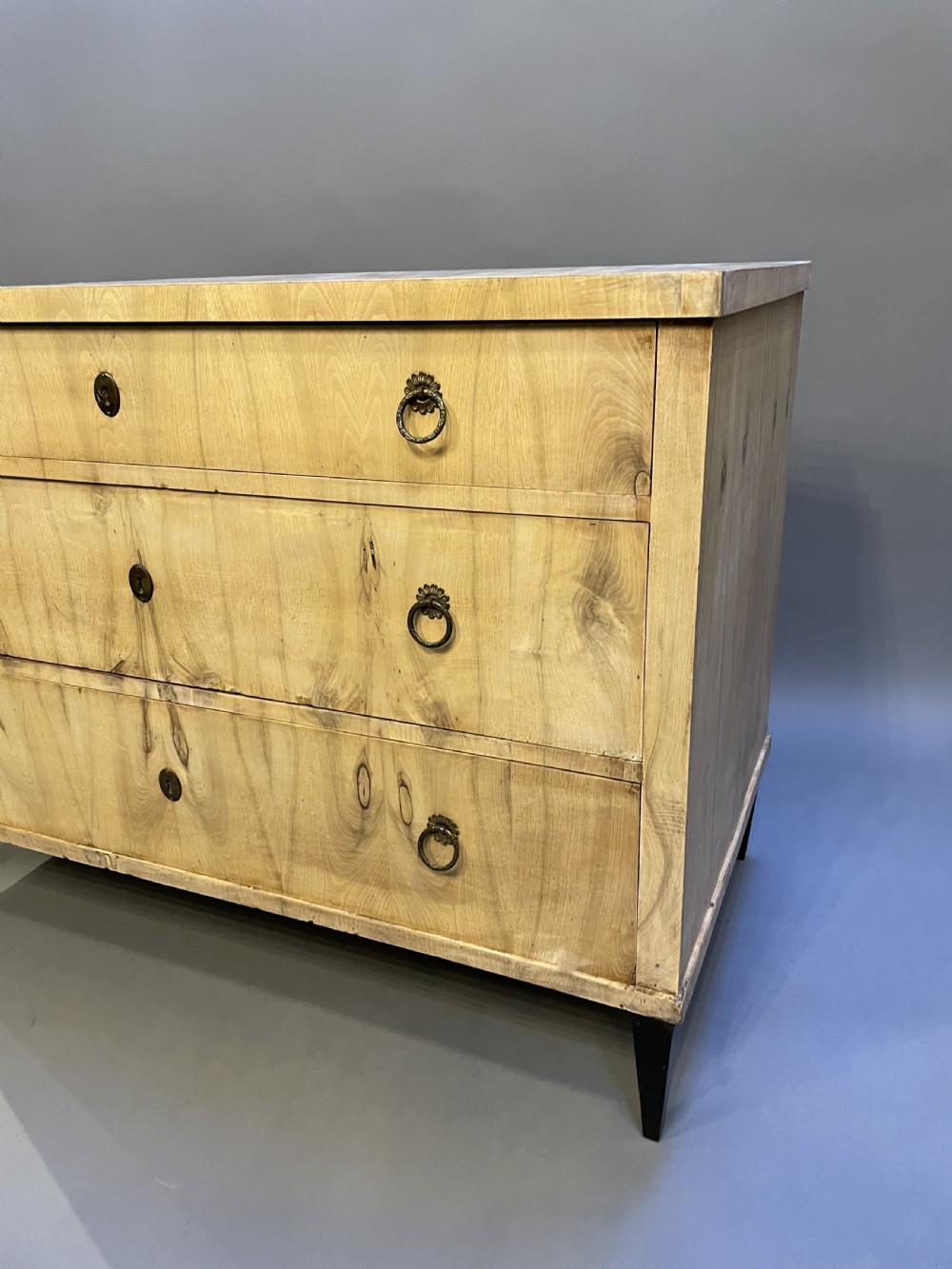 A nineteenth-century Continental light-toned wood commode featuring three wide drawers with brass ring handles and dark tapered feet against a grey background. View II