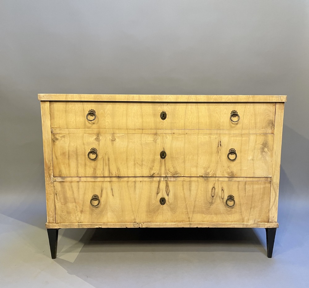 A nineteenth-century Continental light-toned wood commode featuring three wide drawers with brass ring handles and dark tapered feet against a grey background. View III