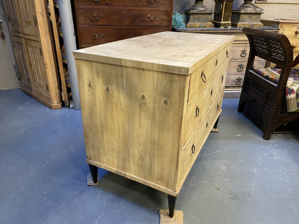A nineteenth-century Continental light-toned wood commode featuring three wide drawers with brass ring handles and dark tapered feet against a grey background. View V