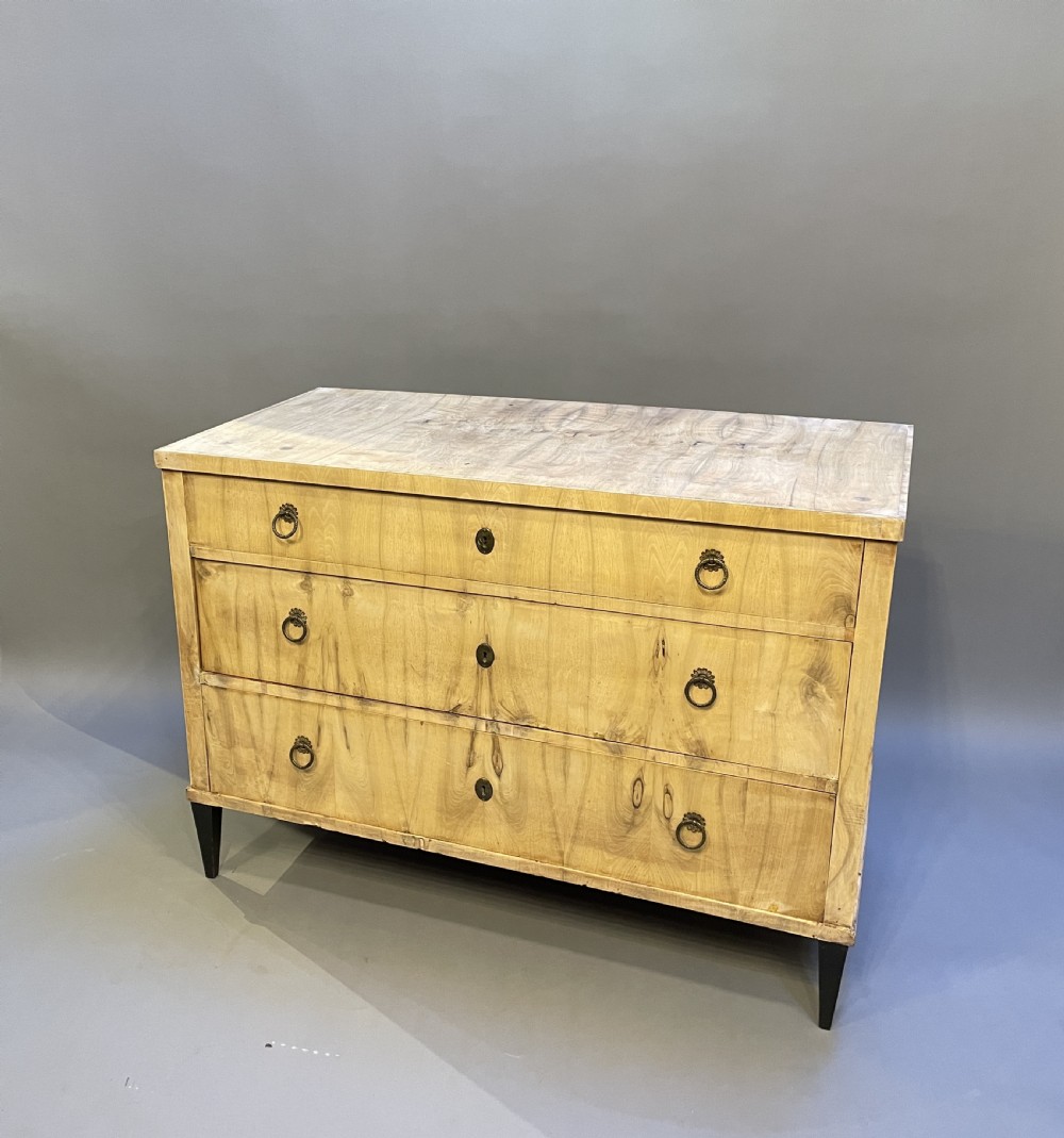 A nineteenth-century Continental light-toned wood commode featuring three wide drawers with brass ring handles and dark tapered feet against a grey background. View VI