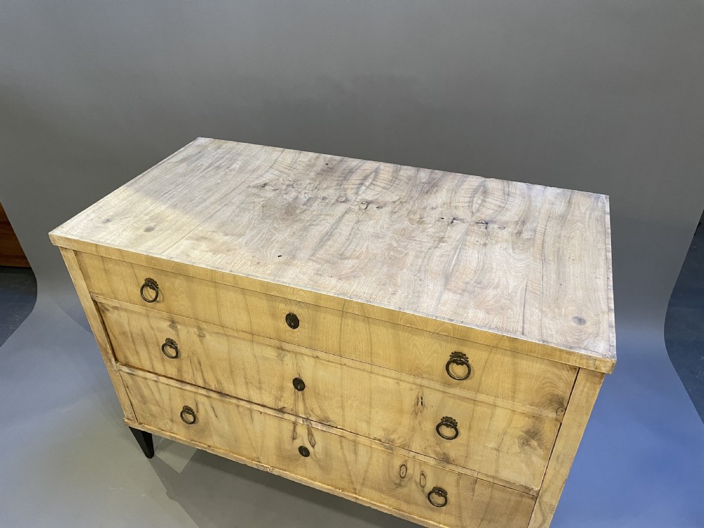 A nineteenth-century Continental light-toned wood commode featuring three wide drawers with brass ring handles and dark tapered feet against a grey background. View VII