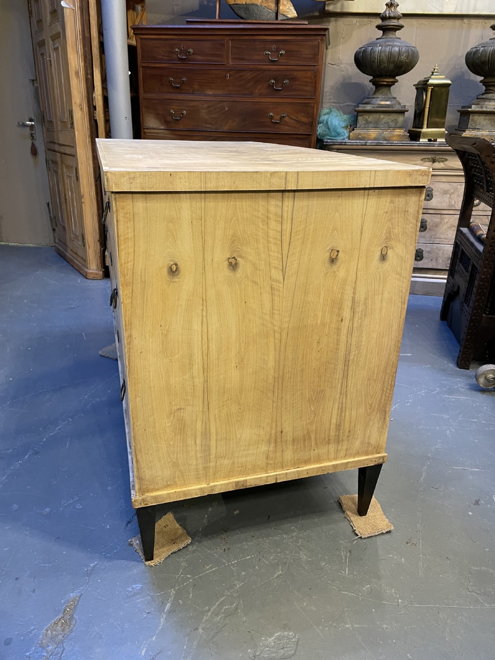 A nineteenth-century Continental light-toned wood commode featuring three wide drawers with brass ring handles and dark tapered feet against a grey background. View VIII