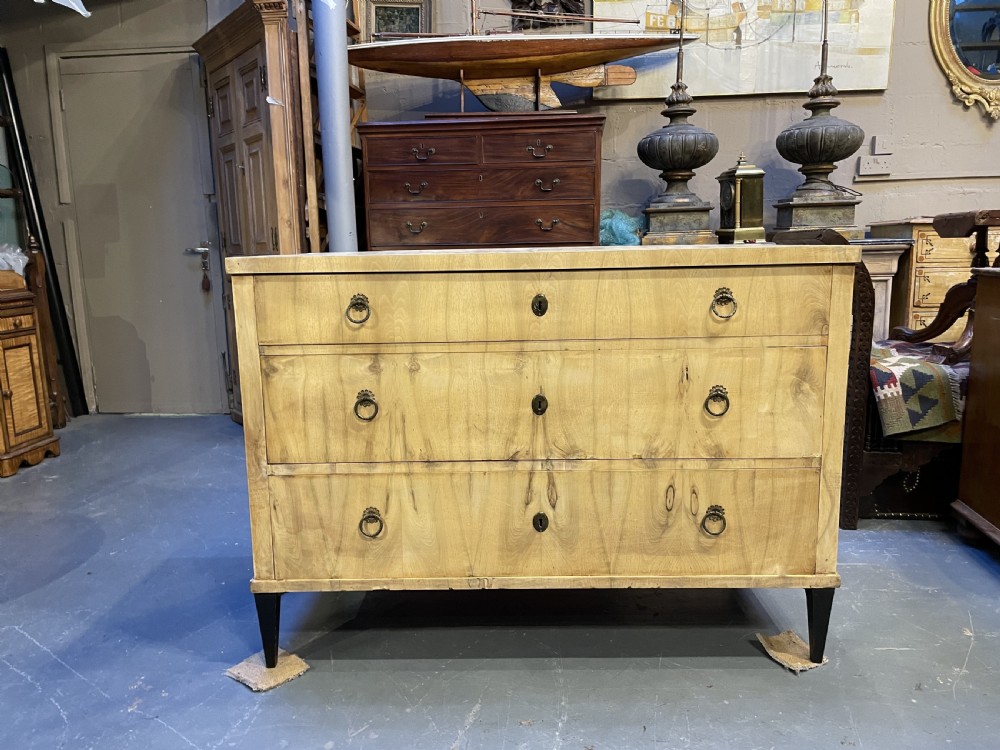 A nineteenth-century Continental light-toned wood commode featuring three wide drawers with brass ring handles and dark tapered feet against a grey background. View X