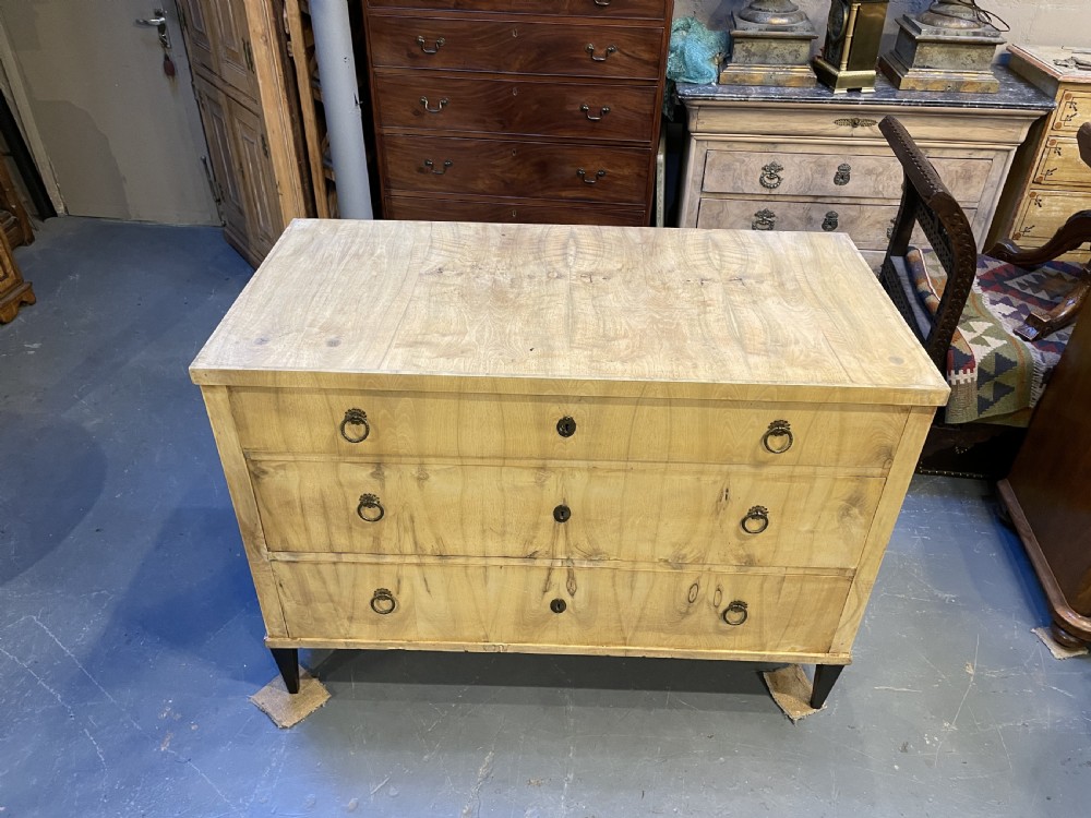A nineteenth-century Continental light-toned wood commode featuring three wide drawers with brass ring handles and dark tapered feet against a grey background. View XI