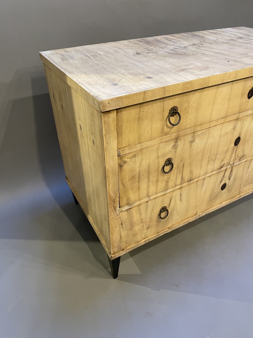 A nineteenth-century Continental light-toned wood commode featuring three wide drawers with brass ring handles and dark tapered feet against a grey background. View XII