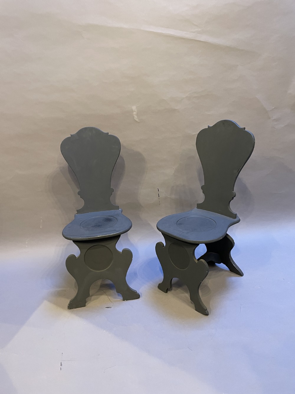 A pair of grey painted wooden Sgabello hall chairs featuring shaped cartouche backs and carved trestle supports.