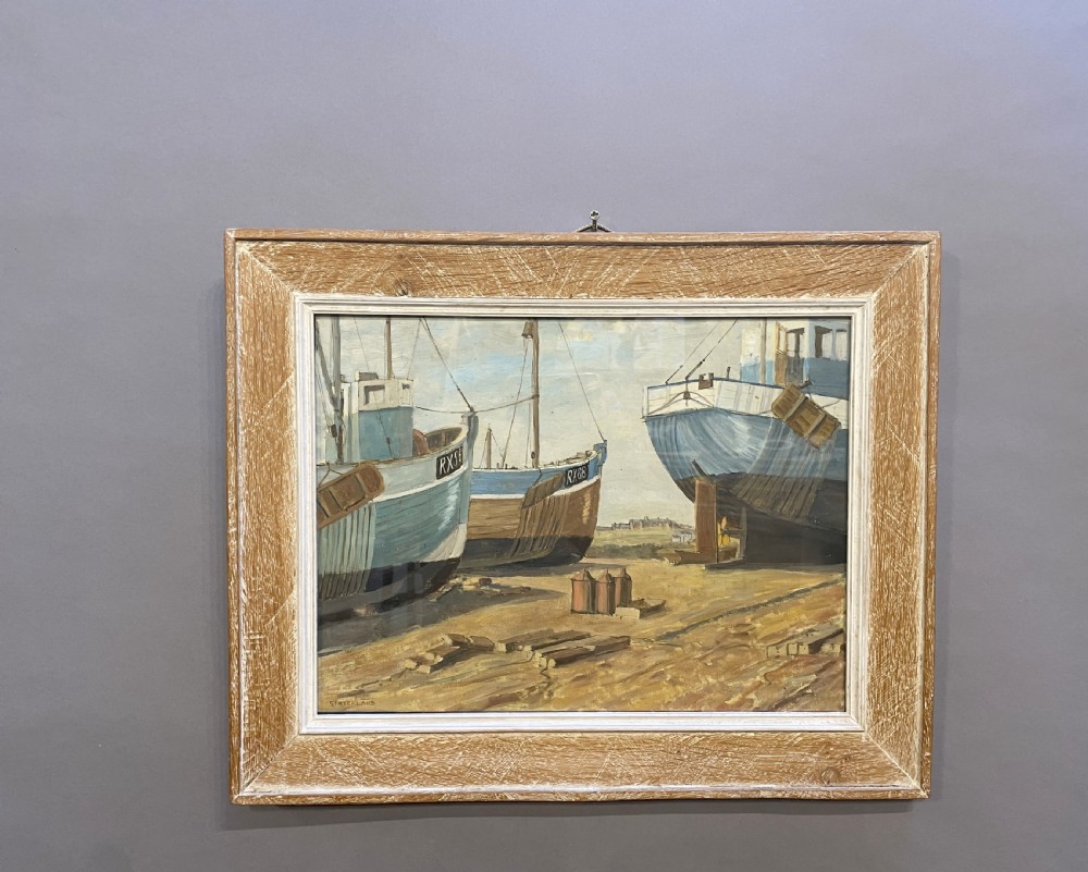 An oil painting on board depicting fishing boats drydocked in a boatyard, framed in light limed wood.