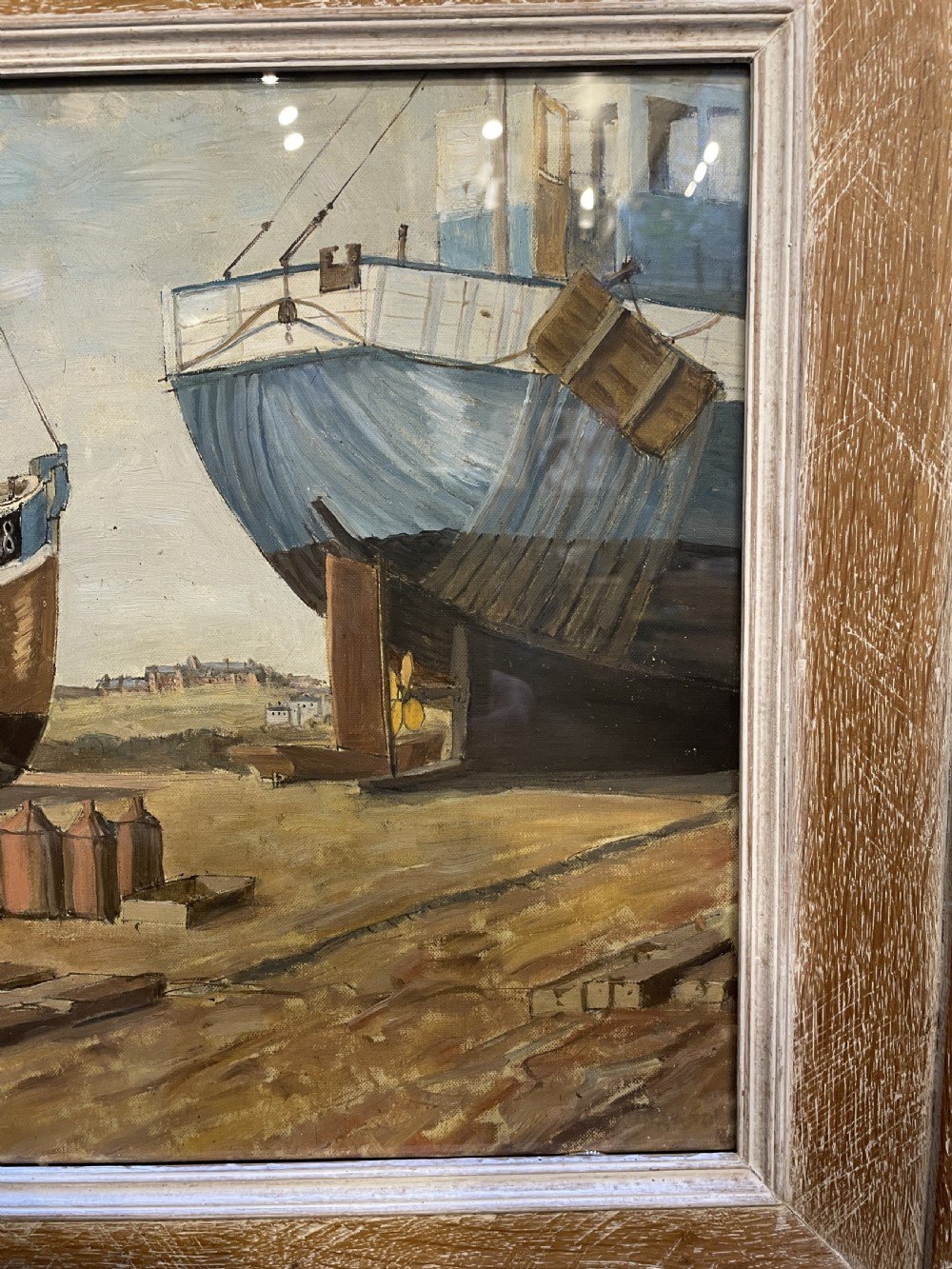 An oil painting on board depicting fishing boats drydocked in a boatyard, framed in light limed wood. View II
