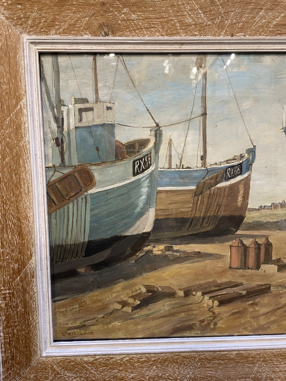 An oil painting on board depicting fishing boats drydocked in a boatyard, framed in light limed wood. View III